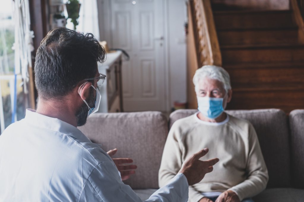 Male professional doctor consulting senior patient during medical care visit wearing masks.